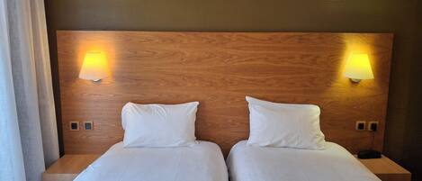Habitación de hotel con dos camas individuales, cabecero de madera, lámparas de noche y una decoración en tonos neutros.