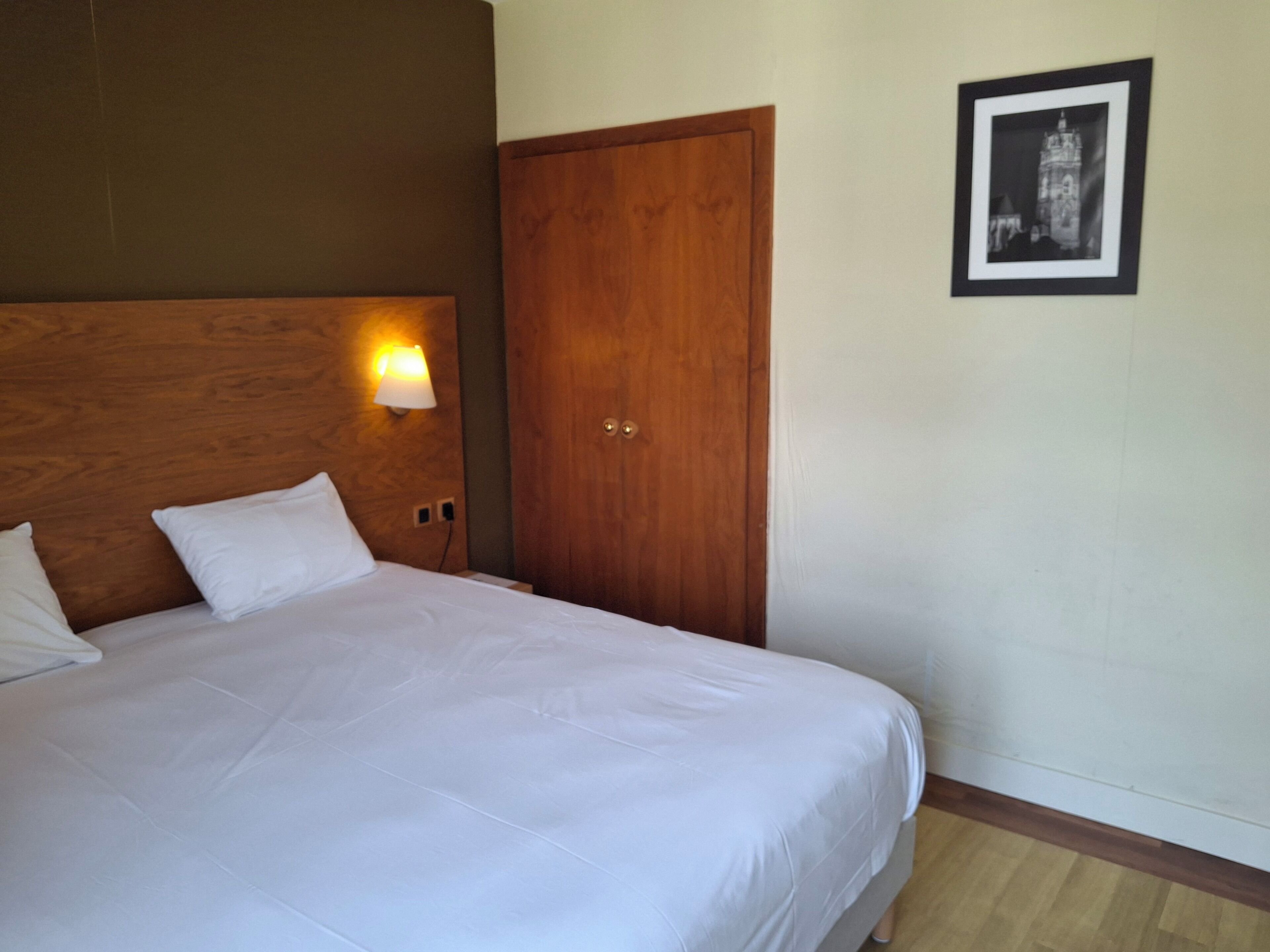 Quarto de hotel com cama, cabeceira de madeira, luminária na parede, porta e quadro na parede.