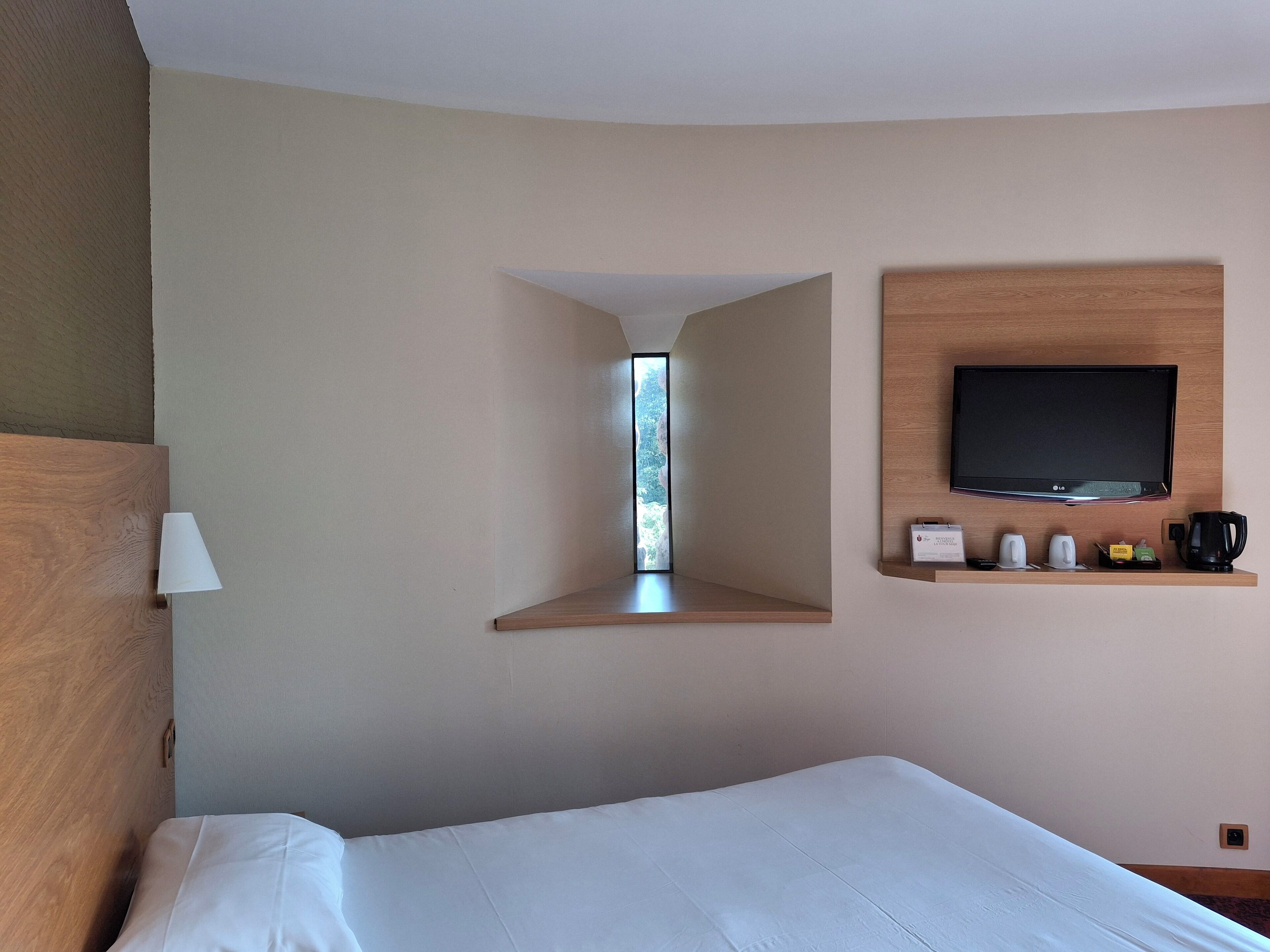 A hotel room with a bed, a mounted TV, a shelf with toiletries, and a window with a view of greenery.