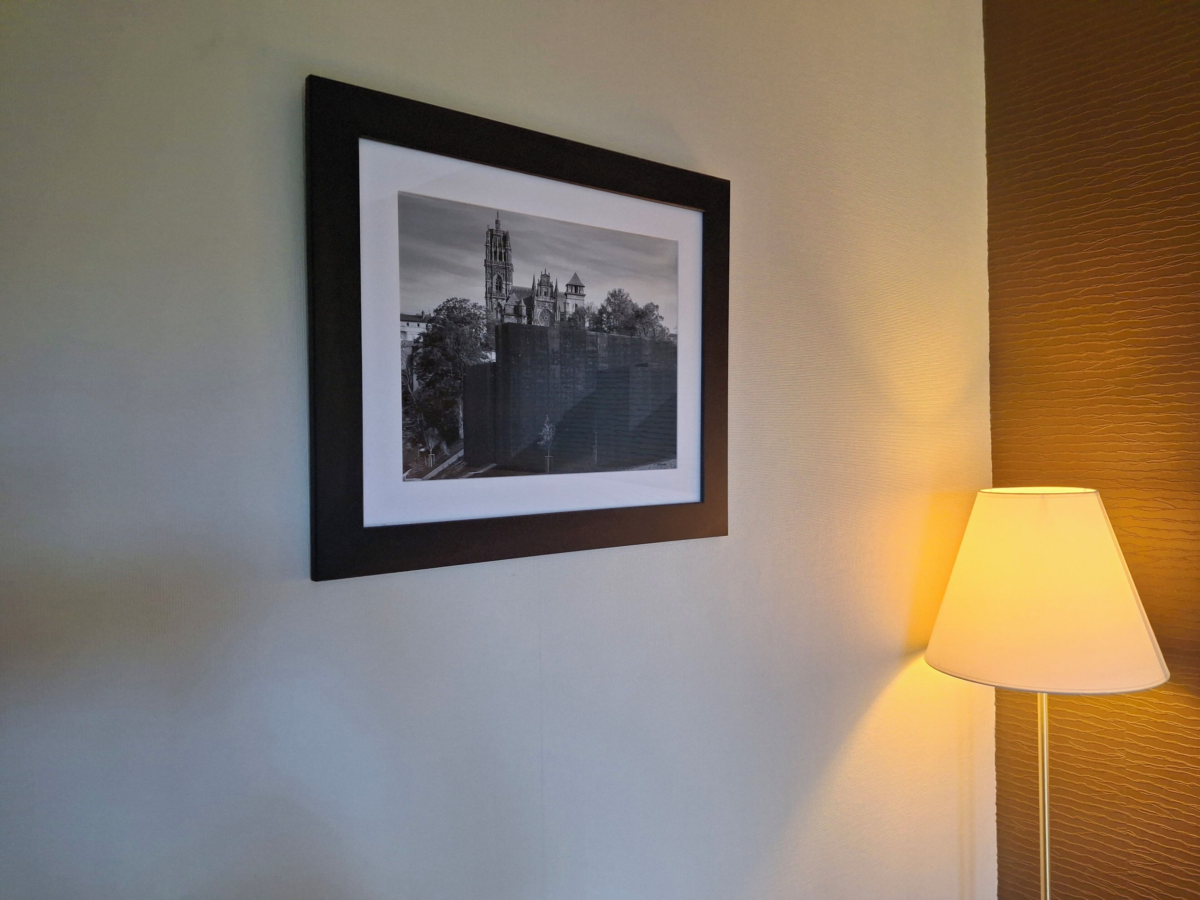 Fotografía en blanco y negro enmarcada de un edificio histórico en una pared, junto a una lámpara de pie.