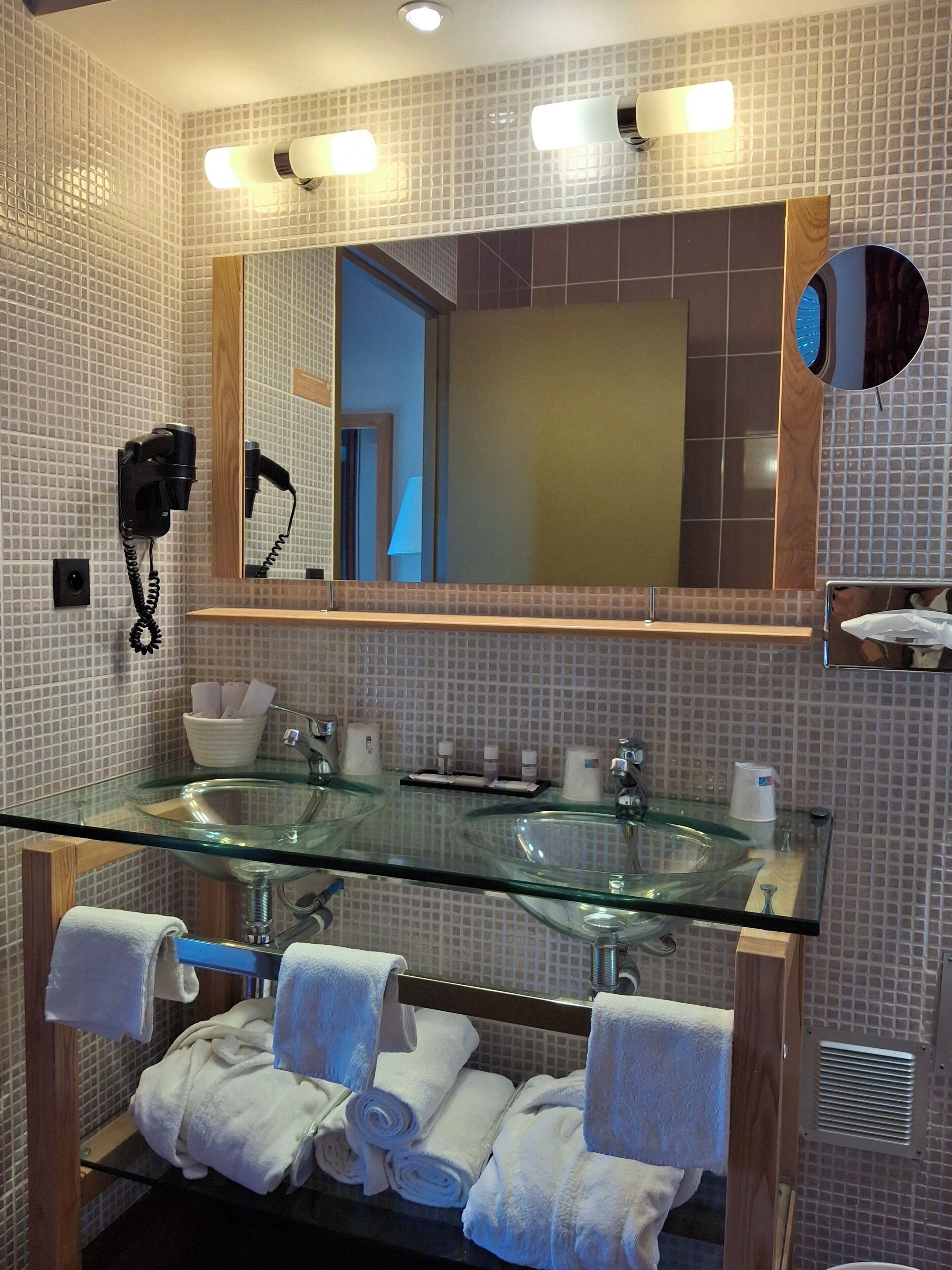 A modern bathroom with a glass sink, a large mirror, and a hairdryer mounted on the wall.