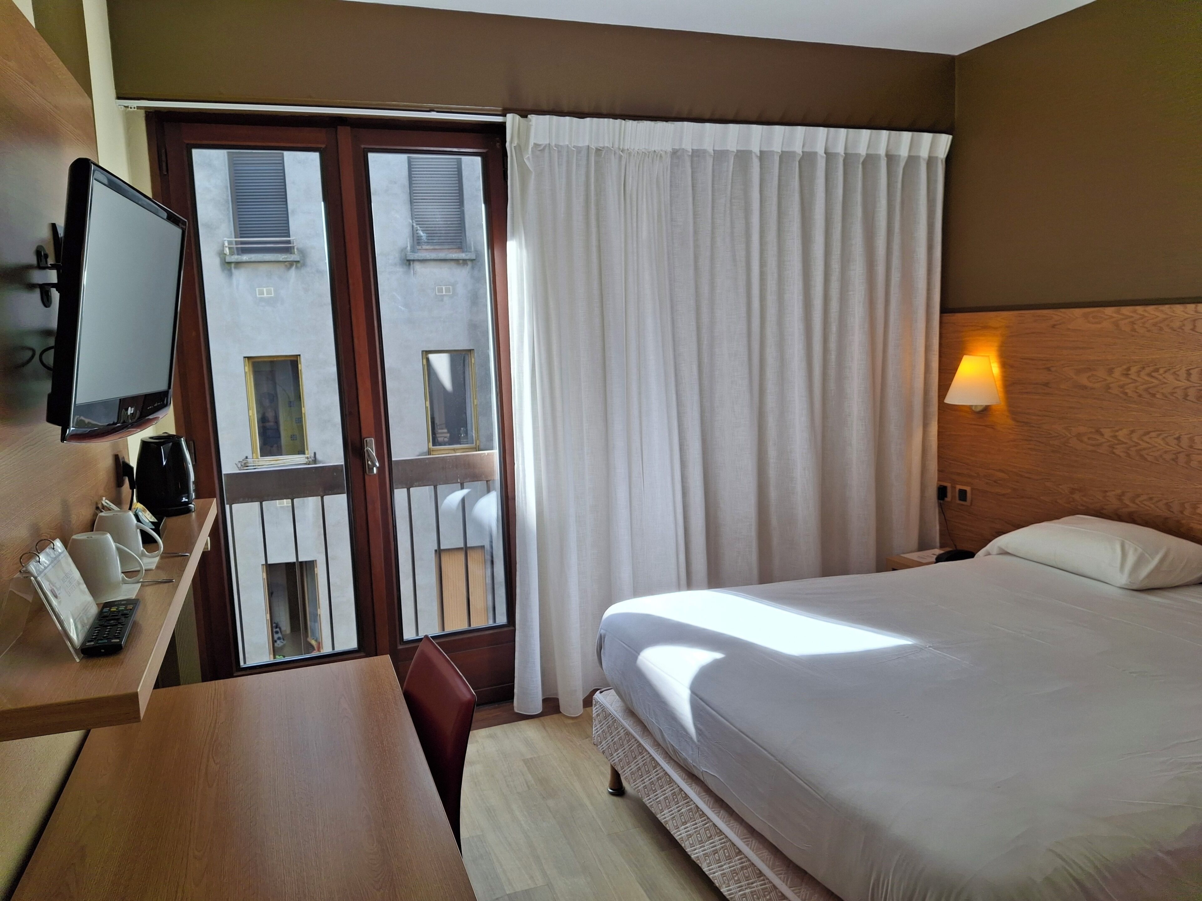 Habitación de hotel con cama, escritorio con monitor de computadora, cafetera y vista al exterior por una puerta de vidrio.