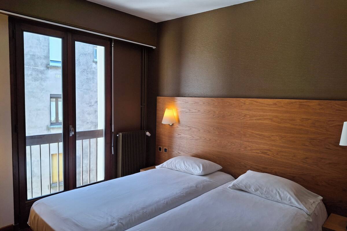 A hotel room with two beds, a wooden headboard, a wall-mounted night light, and a sliding glass door leading to an outdoor balcony.