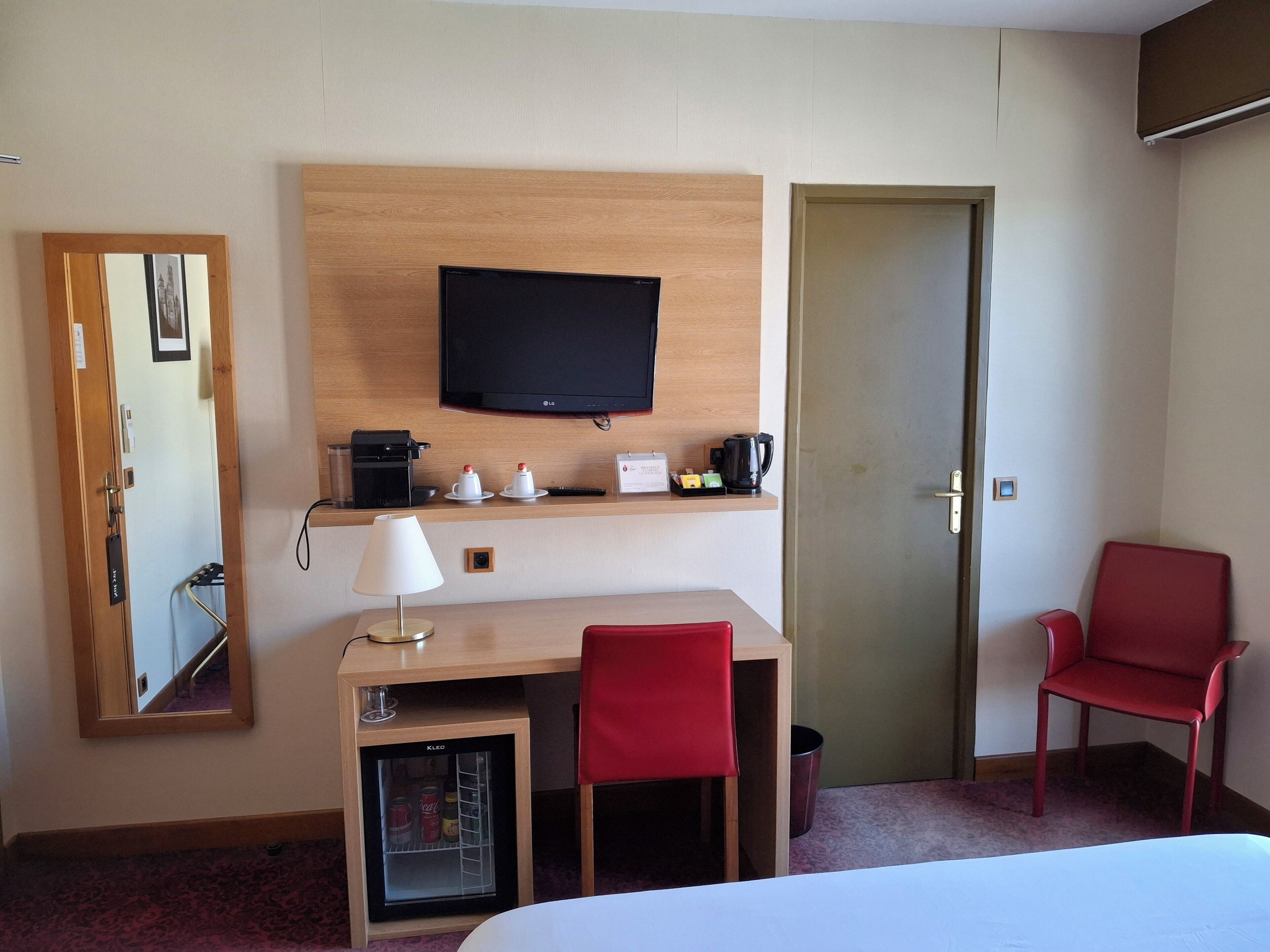 Une chambre d’hôtel comprenant un lit, un bureau avec une chaise, une télévision, un mini-réfrigérateur, une machine à café et une chaise rouge.