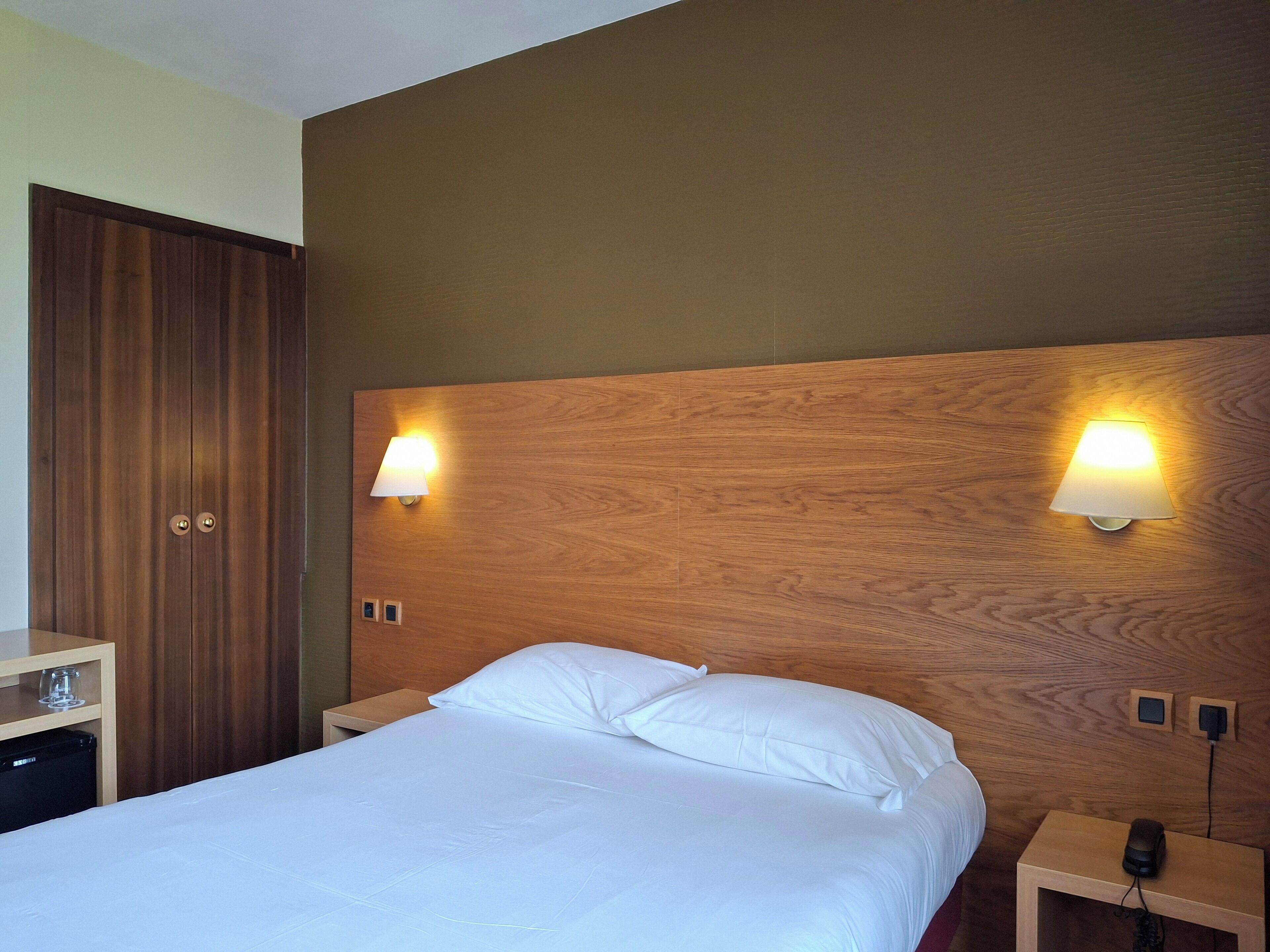 Habitación de hotel con cabecera de madera, dos lámparas de noche, cama con ropa de cama blanca, mesita de noche con un teléfono y un cargador, y armario con espejo.