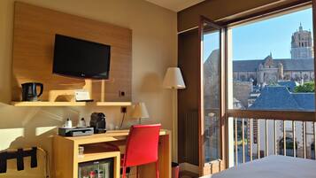 Habitación de hotel con cama, escritorio y silla, televisor, cafetera, refrigerador pequeño y ventana con vistas a una catedral.