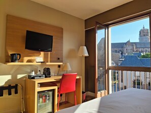 Une chambre d’hôtel comprenant un lit, un bureau avec une chaise, une télévision, une machine à café, un mini-réfrigérateur et une fenêtre donnant sur une cathédrale.