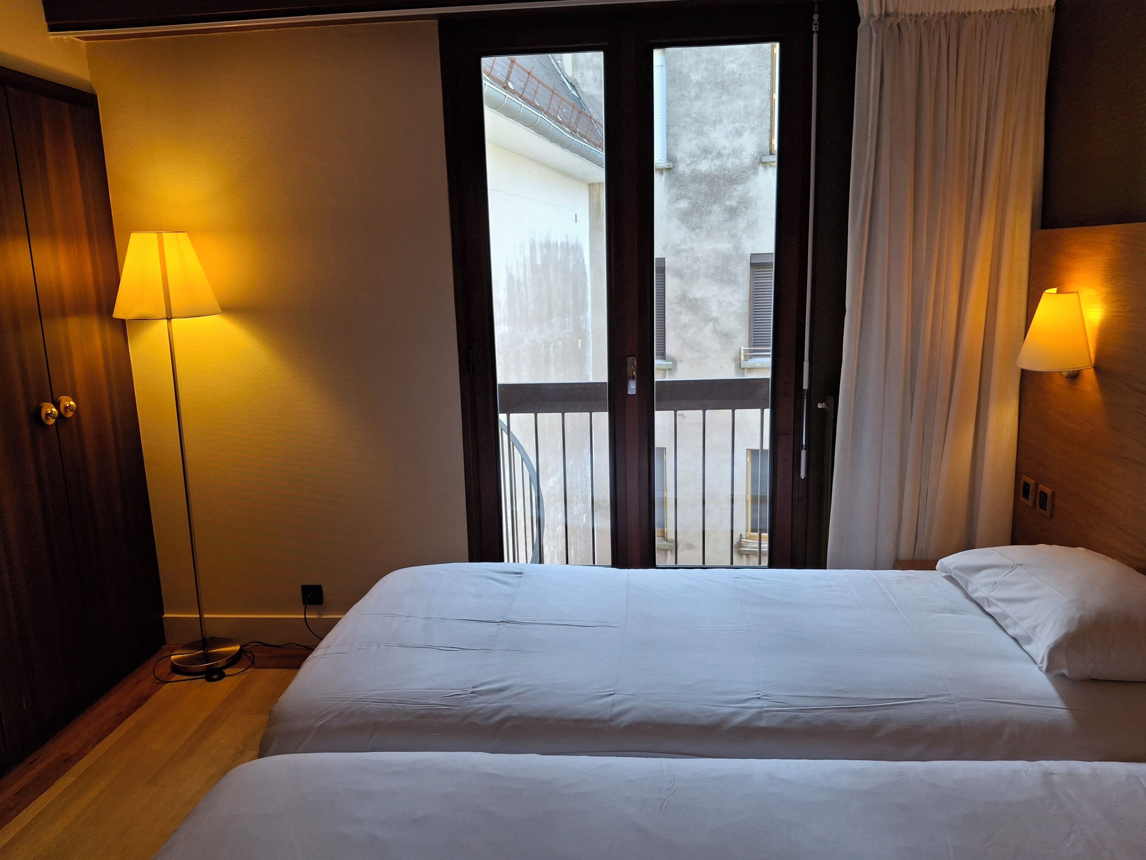 A hotel room with a bed, two bedside lamps, a wooden wardrobe, and a view through a glass door.