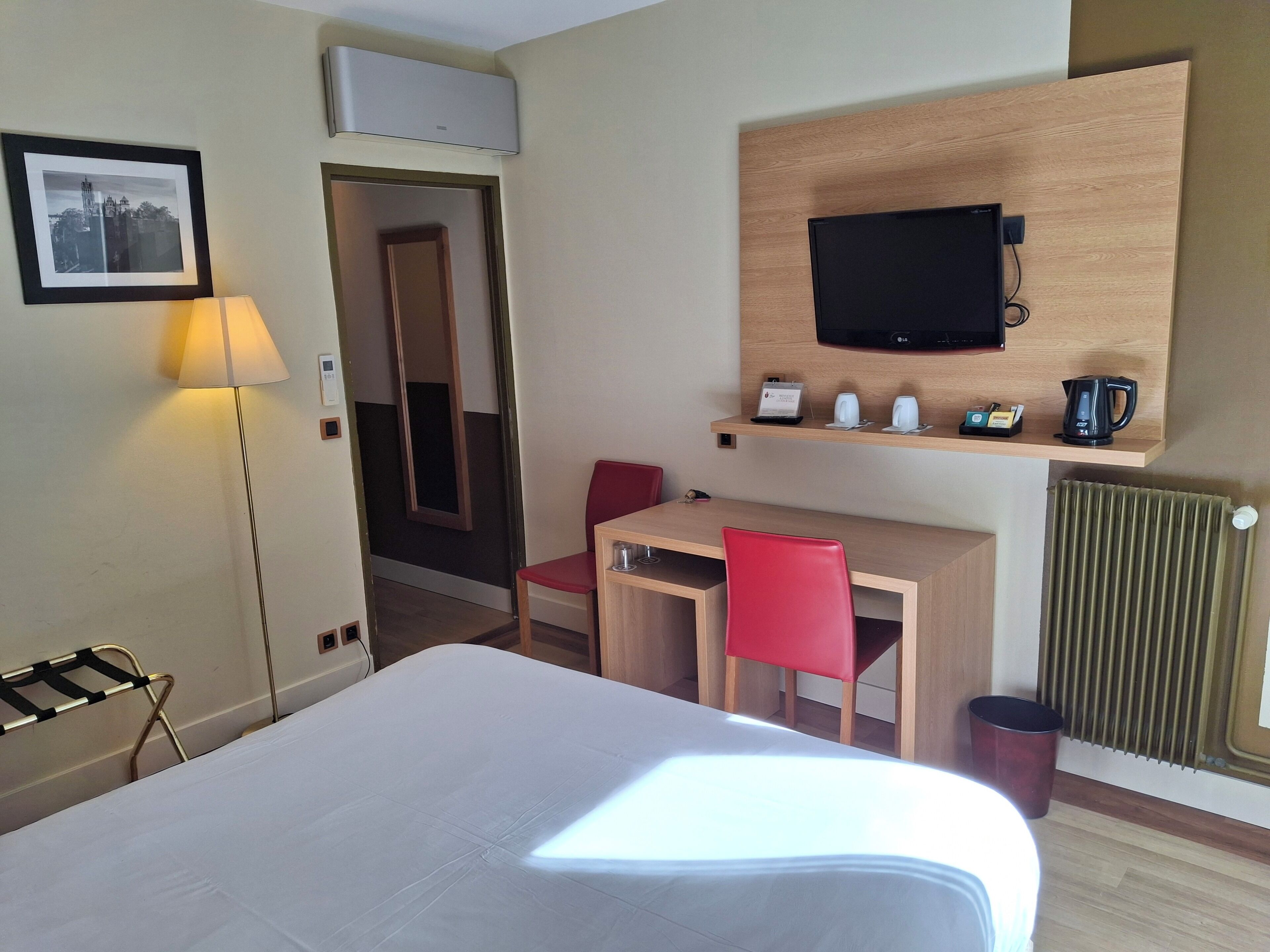 A hotel room with a bed, desk, red chair, TV, and a small shelf with a kettle and cups.