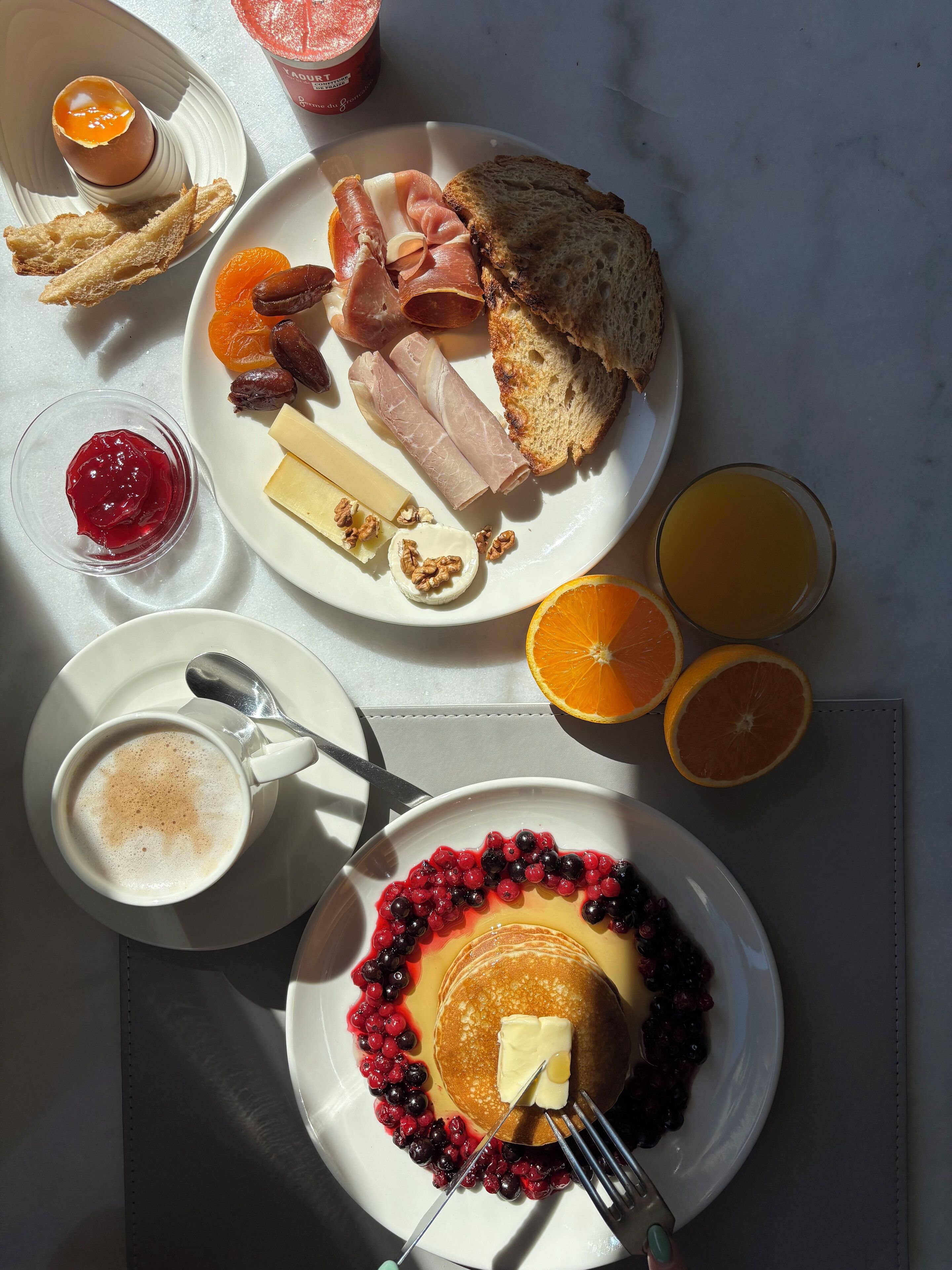 Una mesa de desayuno con platos de comida, incluyendo tostadas, carnes, queso y frutas, además de una taza de café y un vaso de jugo de naranja.