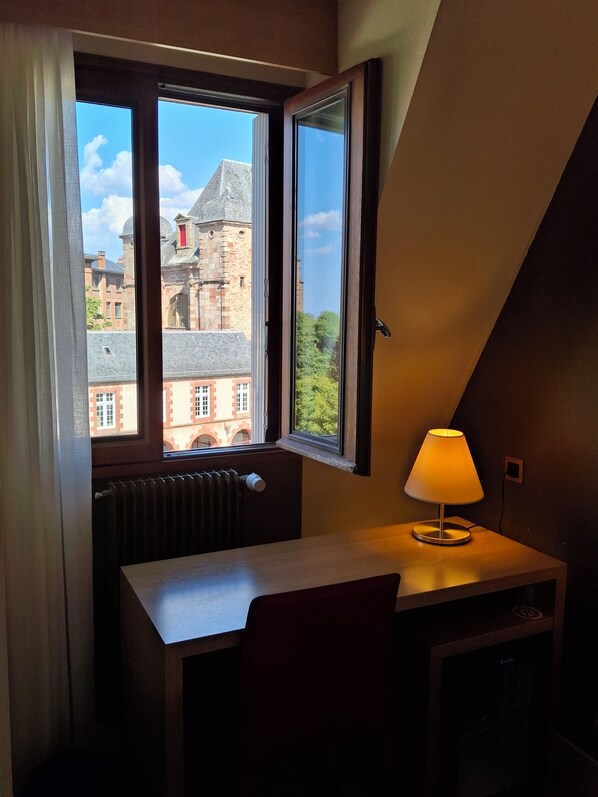 A room with a window overlooking a historic building, a radiator, a desk with a lamp, and a chair. - Hôtel Tour Maje (Rodez)