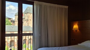 A hotel room with a bed, a window with curtains, a view of a historic building, and a wall-mounted lamp. - Hôtel La Tour Maje (Rodez)