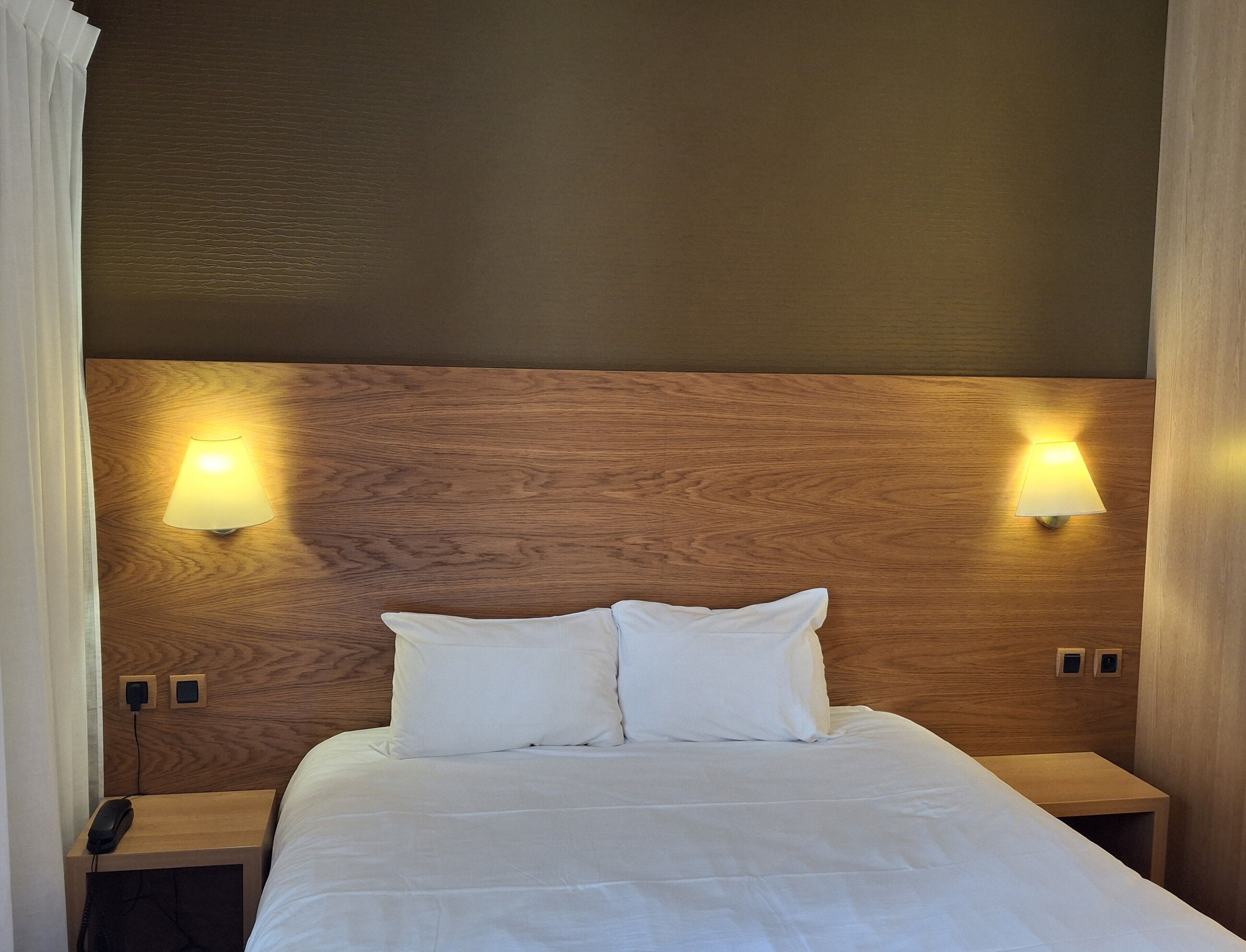 A hotel room with a wooden headboard, two bedside lamps, a bed with white bedding, and a bedside telephone.