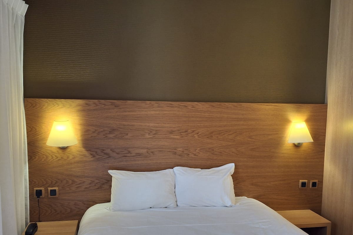 Una habitación de hotel con cabecera de madera, dos lámparas de noche, una cama con ropa de cama blanca y un teléfono junto a la cama.