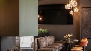 A modern living room with a sofa, a round table, and a chair. There is a fireplace with logs, a plant, and a TV mounted on the wall. - Hôtel & Spa Le Maury, The Originals Boutique, Vannes (Vannes)
