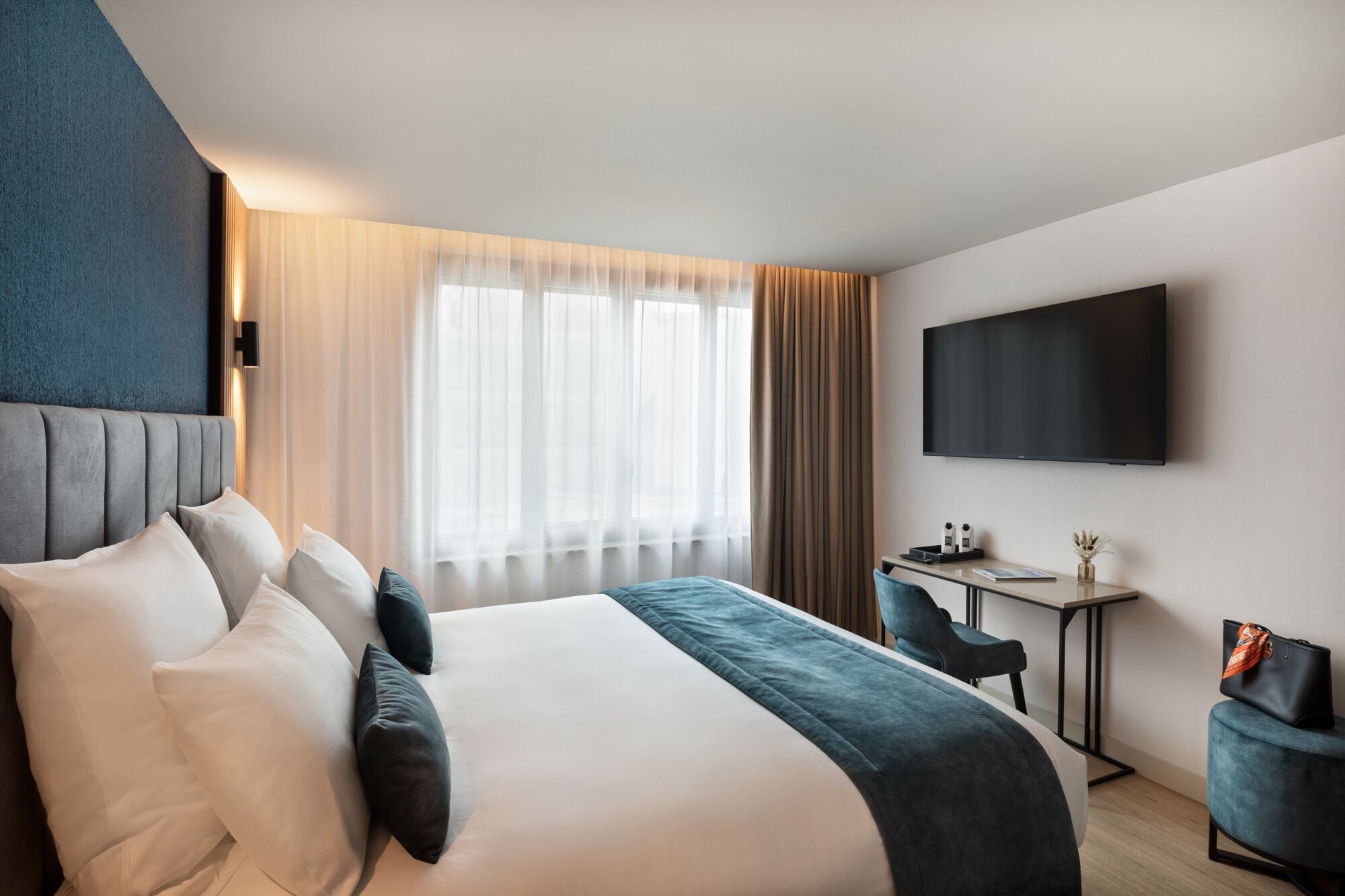 A hotel room with a large bed, a desk with a chair, a flat-screen TV, and a window with curtains.