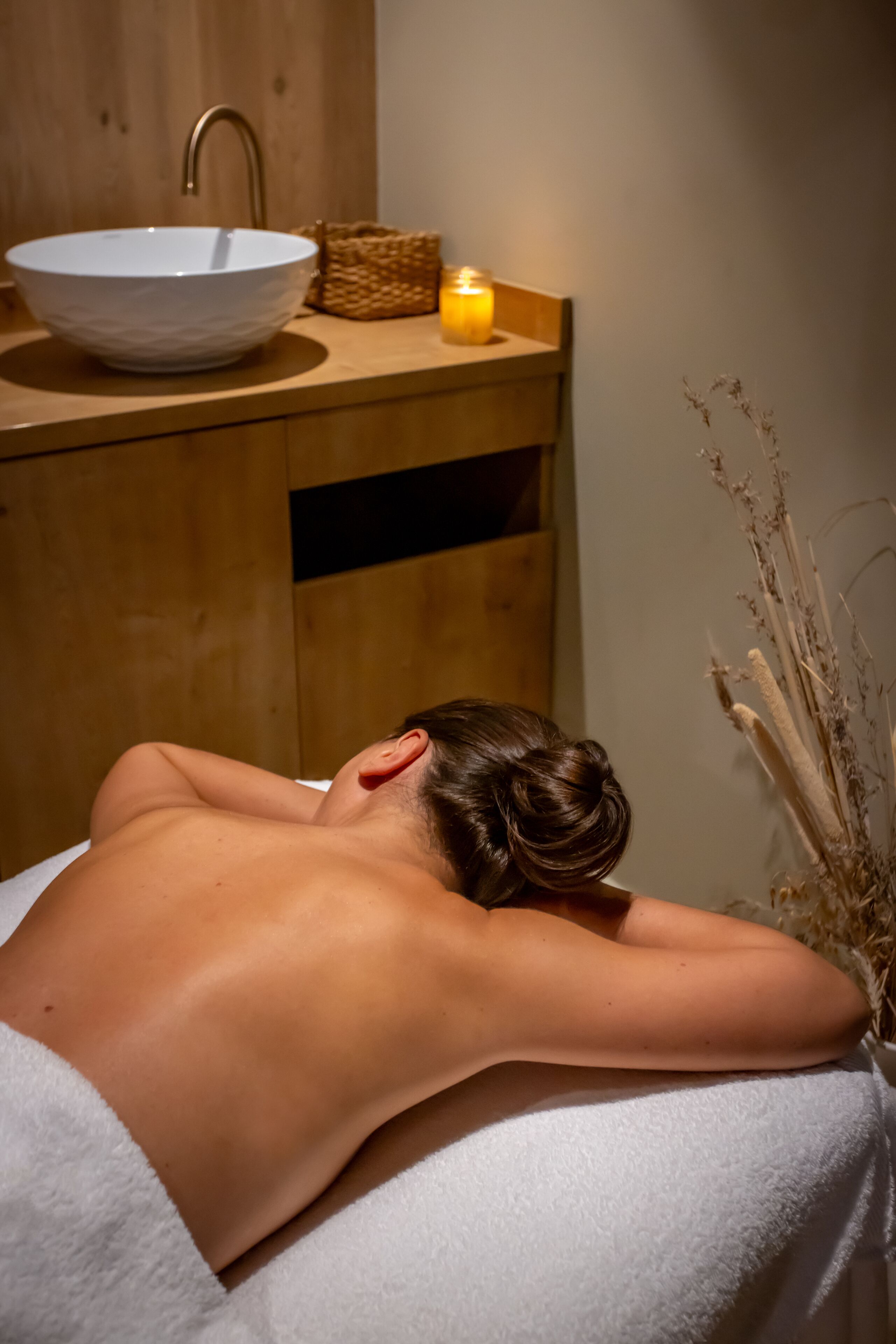 Une personne reçoit un massage du dos dans une pièce où se trouvent un meuble en bois, un lavabo blanc et une bougie.