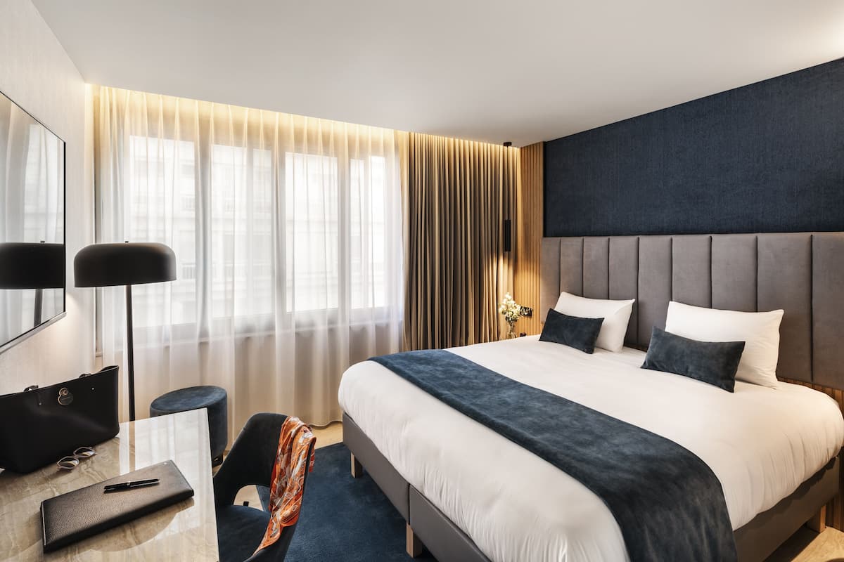 A modern hotel room with a large bed, a desk with a chair, a flat-screen TV, and a large window with curtains.