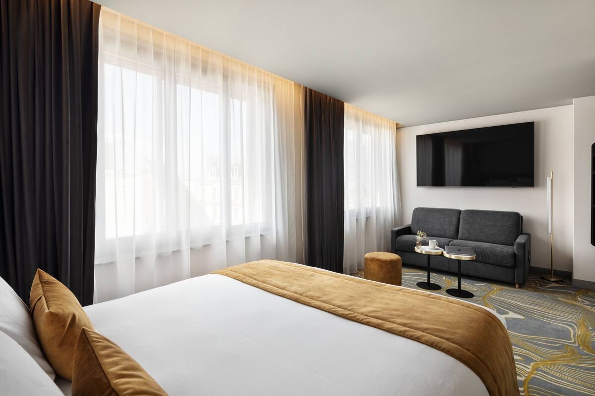 A hotel room with a large bed, a sofa, a TV mounted on the wall, and a small table with a laptop and a cup.