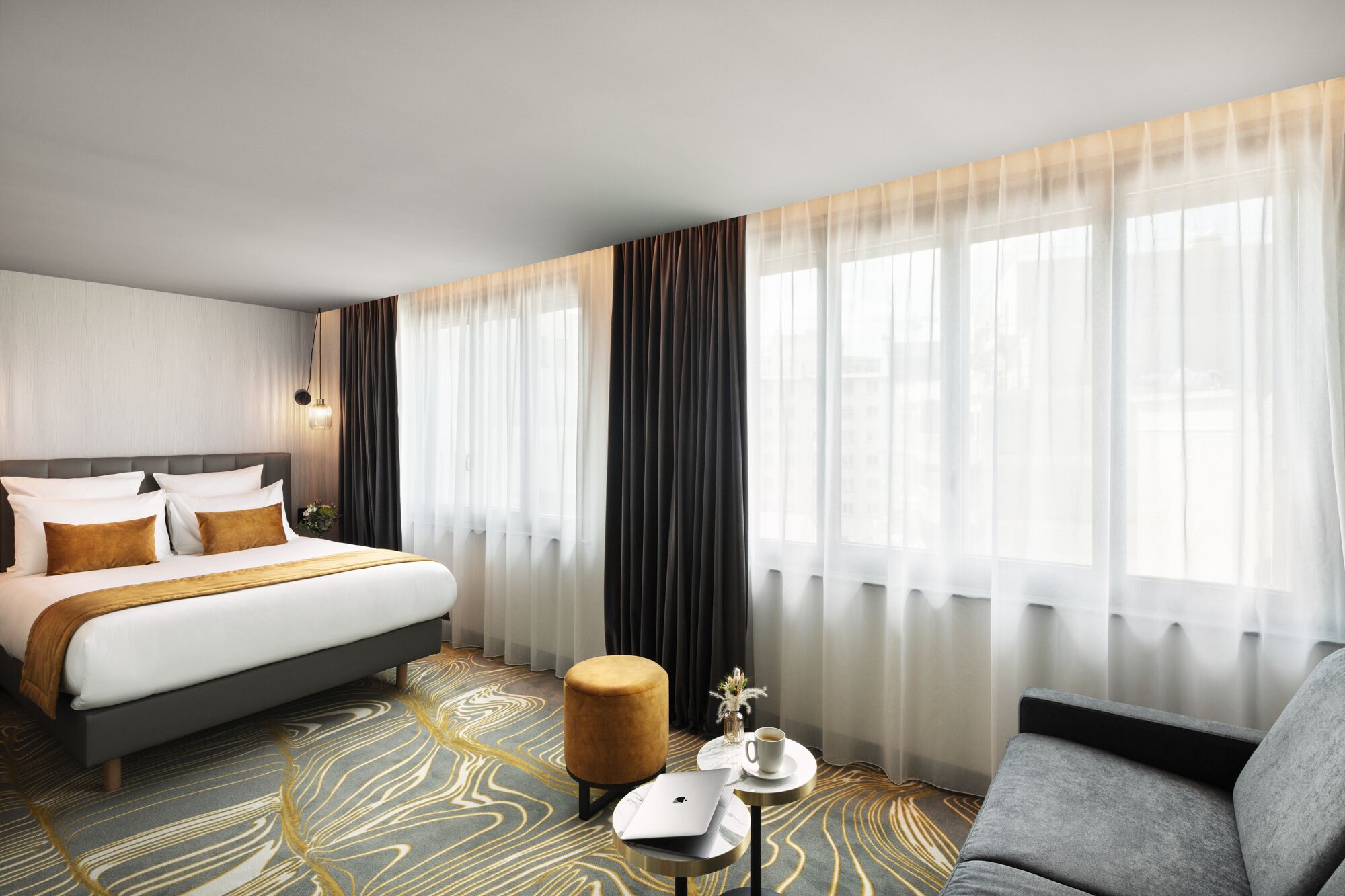 A modern hotel room with a large bed, a sofa, a small table with a cup and a laptop, and a view of the city through sheer curtains.