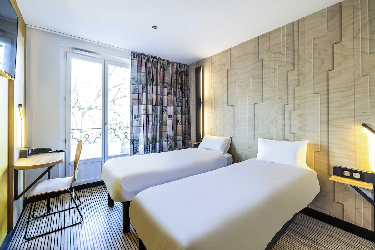 A hotel room with two beds, a desk, a chair, and a view of the outdoors.