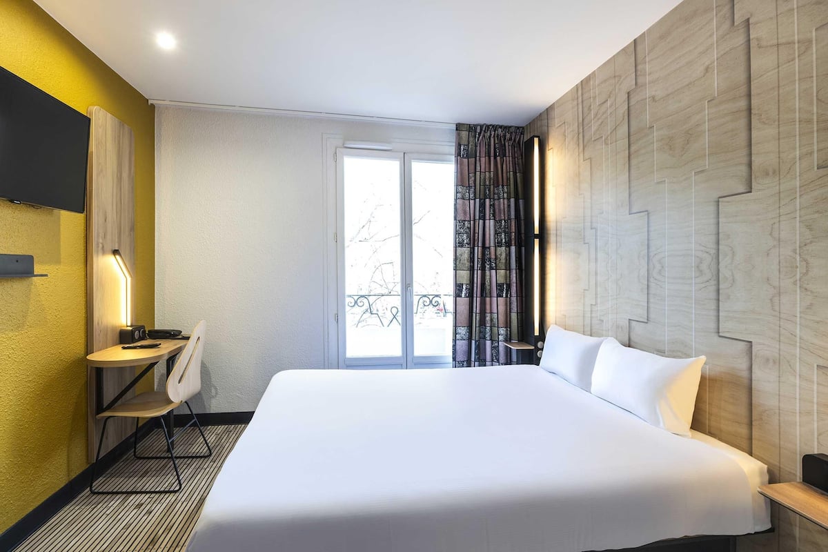 A hotel room with a large bed, a desk with a chair, a TV mounted on the wall, and a window with curtains.