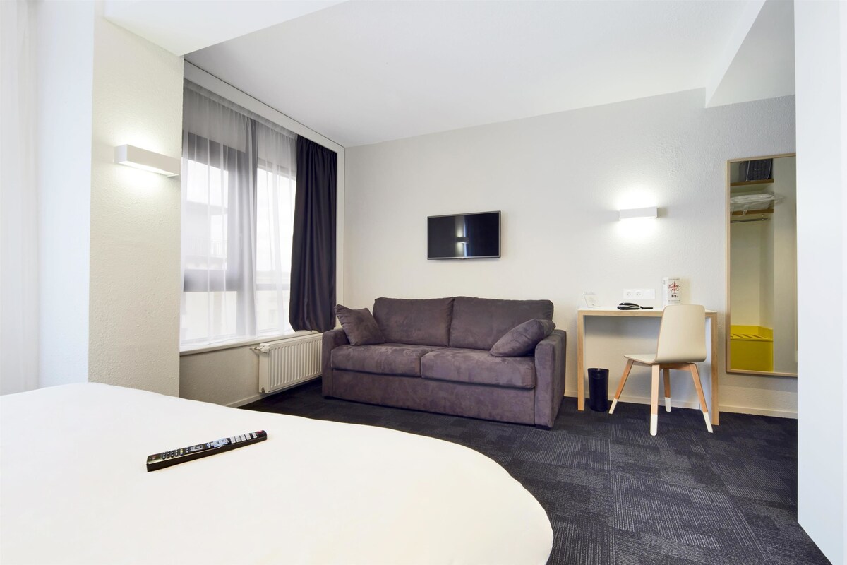 A modern hotel room with a grey sofa, a desk with a chair, a flat-screen TV, and a large mirror.