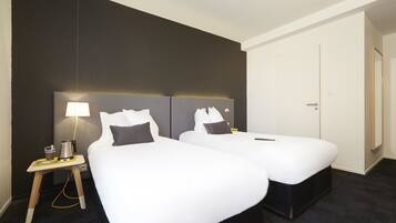 A hotel room with two beds, a bedside table with a lamp, and a door.