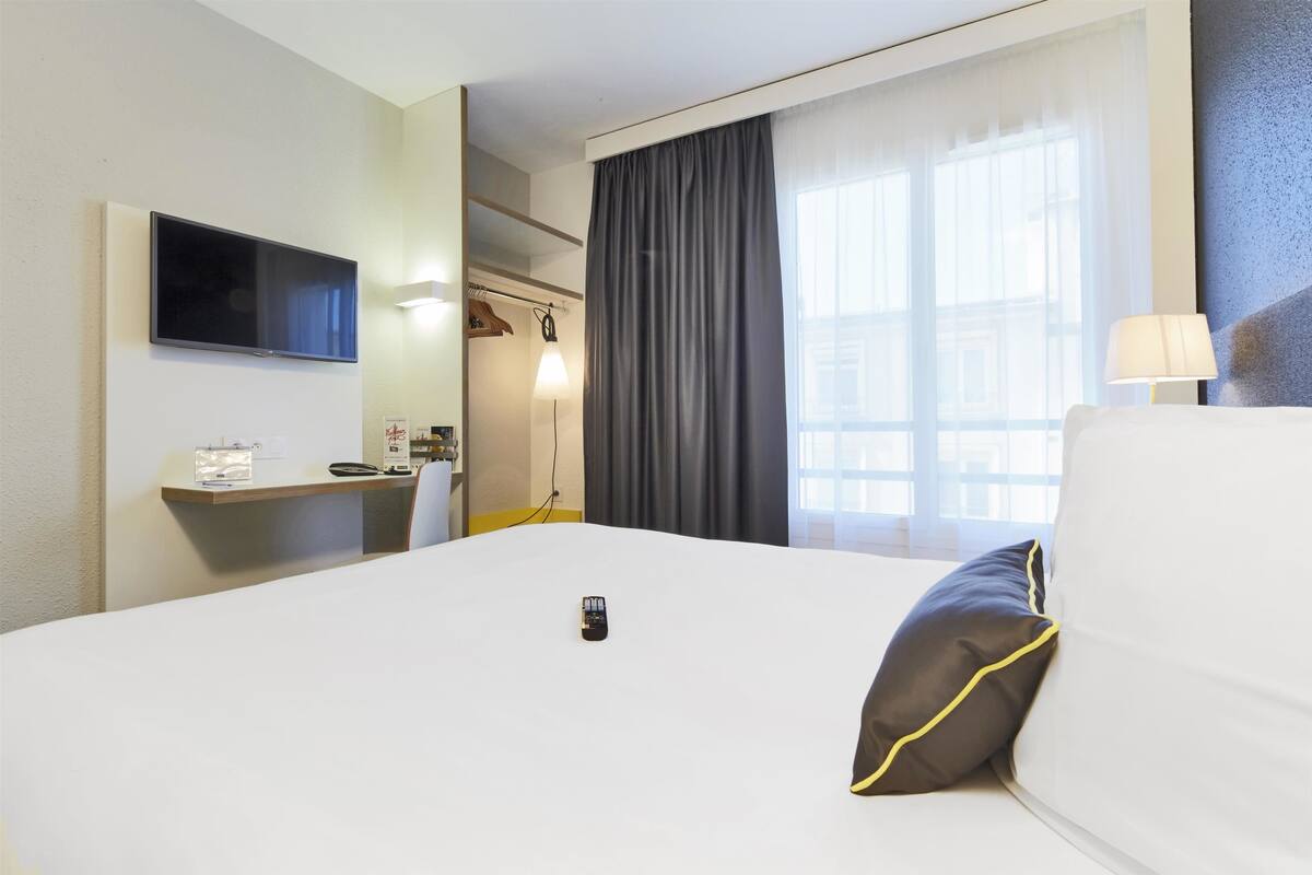 Una camera d'albergo con un letto, una TV a parete, una scrivania con lampada e una finestra con tende.