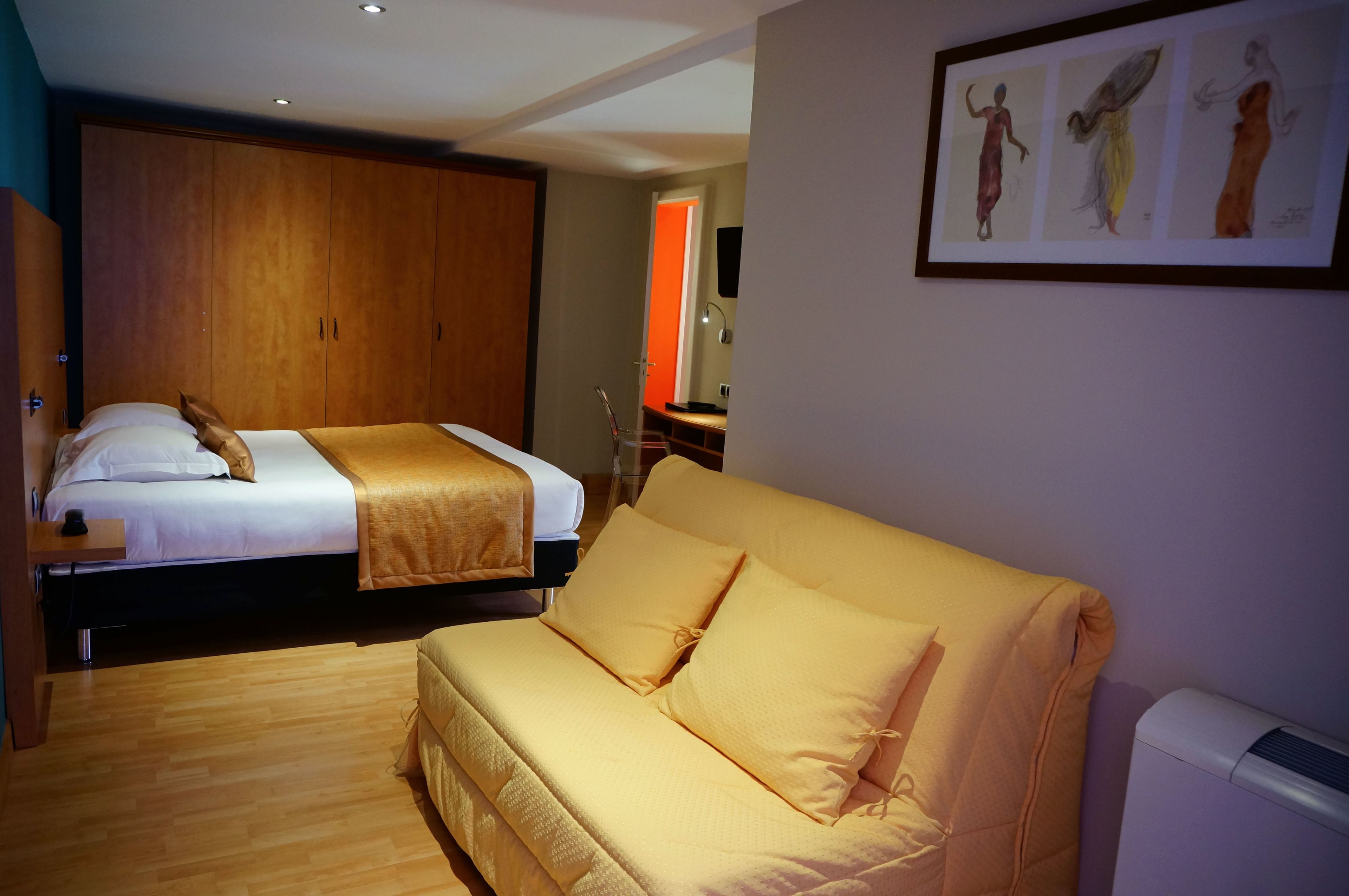 A hotel room with a double bed, a sofa, a wooden wardrobe, and a framed artwork on the wall.