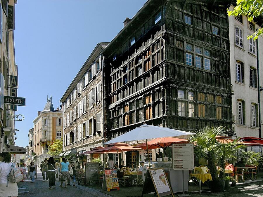 A street with a building that has multiple stories and a traditional facade. There are outdoor seating areas with umbrellas and tables, and a signboard is visible.