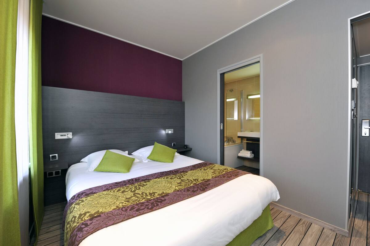 A hotel room with a bed, green pillows, a patterned bedspread, a dark headboard, a mirror, and a bathroom visible through an open door.