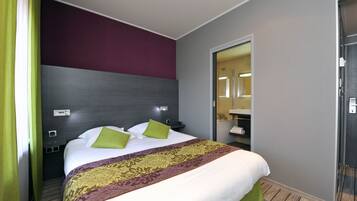 A hotel room with a bed, green pillows, a patterned bedspread, a dark headboard, a mirror, and a bathroom visible through an open door.
