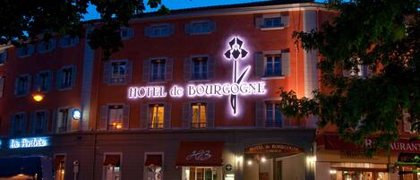 The exterior of Hotel de Bourgogne with illuminated signage, a visible entrance, and surrounding greenery.