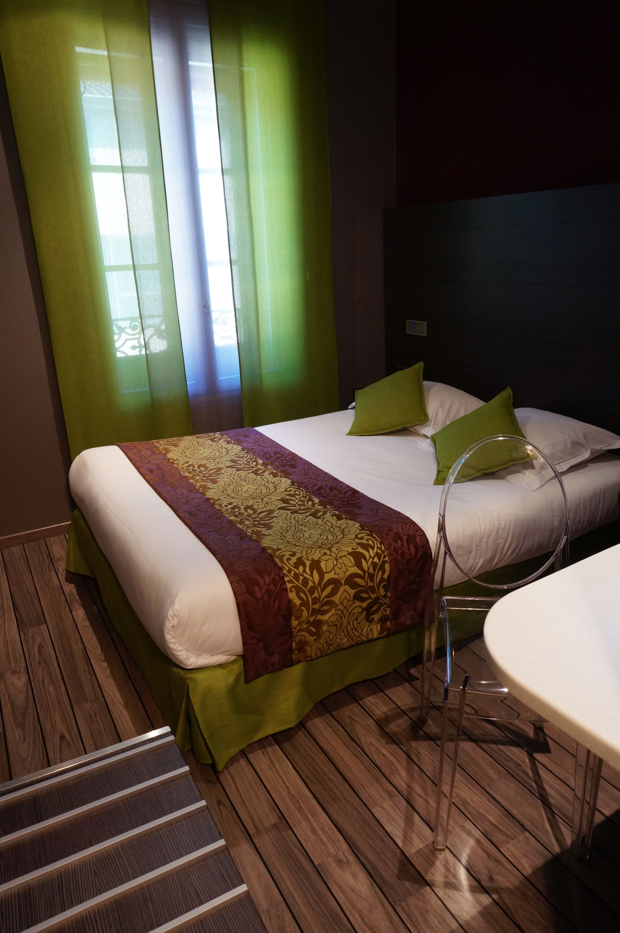 A hotel room with a bed, green curtains, a wooden floor, and a round table.