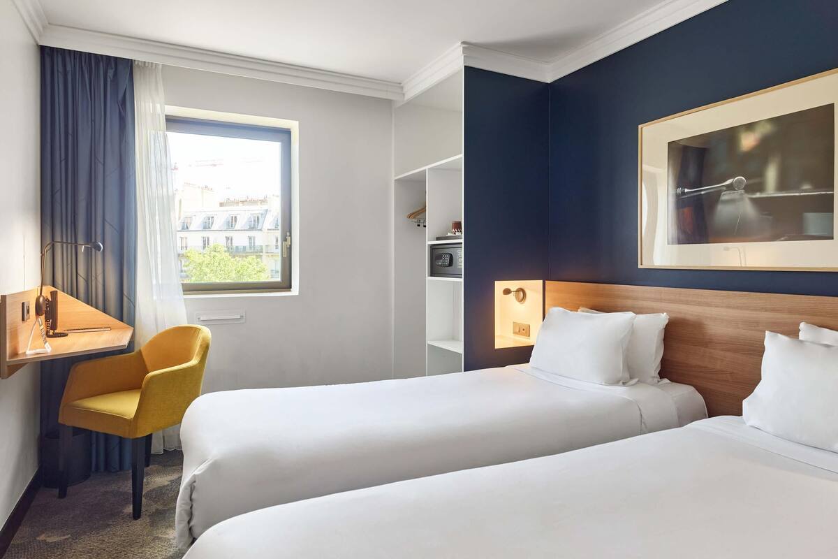 Hotellihuone, jossa on kaksi sänkyä, työpöytä, tuoli, ikkuna, jossa on verhot, seinällä oleva taulu ja huoneen nurkassa mikroaaltouuni.