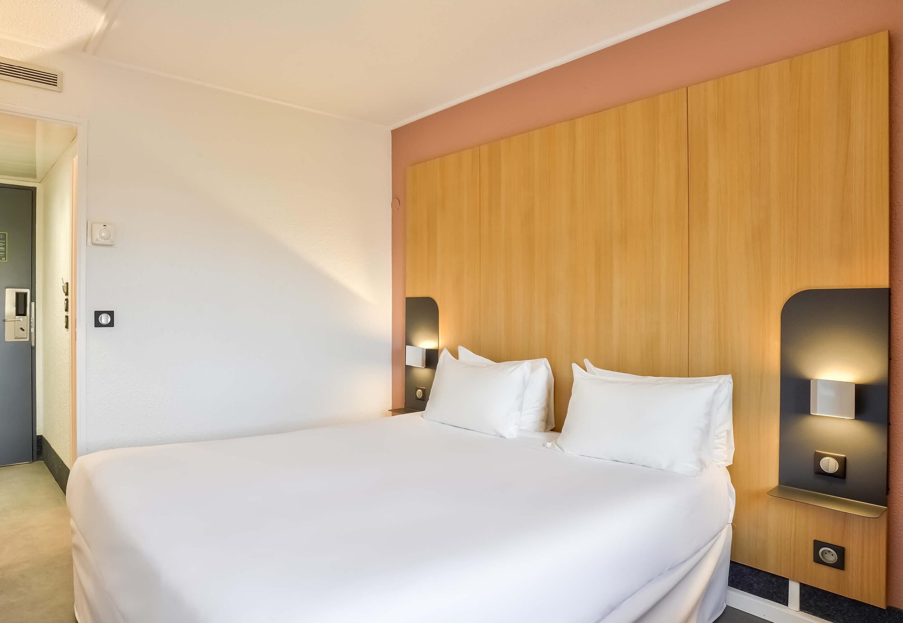 Une chambre d’hôtel avec un grand lit, une tête de lit en bois, des lampes de chevet et une porte donnant sur une autre pièce.