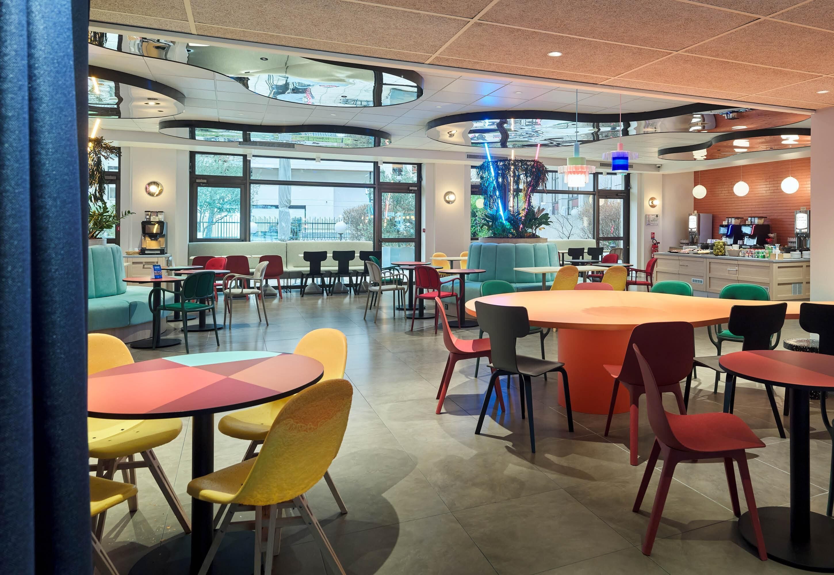 Un espace de restauration intérieur moderne, doté de tables rondes et de chaises colorées, de grandes fenêtres et d’un coin salon central avec un canapé bleu.