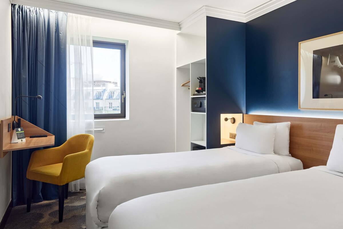 Hotellihuone, jossa on kaksi sänkyä, työpöytä, tuoli, vaatekaappi ja ikkuna, jossa on verhot.