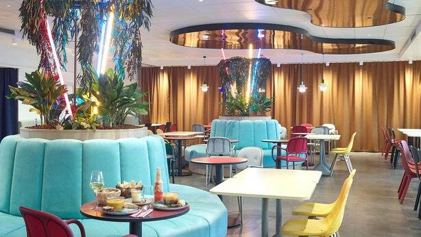 Ein Restaurant mit runden Tischen, blauen und roten Stühlen und einer zentralen Pflanzenausstellung.