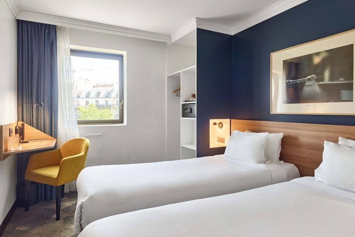 Habitación de hotel con dos camas, un escritorio, una silla, una ventana con cortinas y un cuadro en la pared.