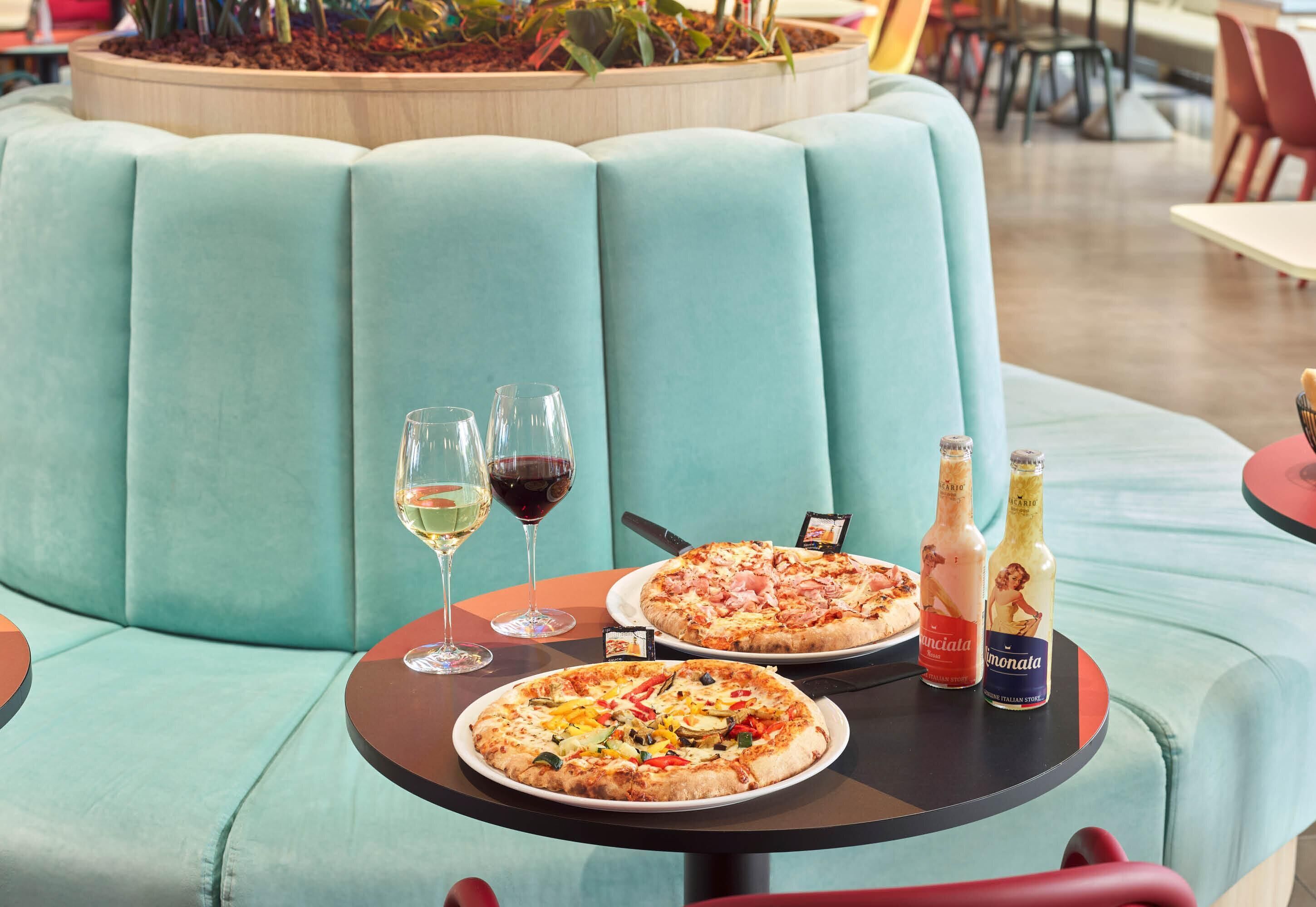 A dining area with a teal cushioned bench, two pizzas on plates, wine glasses, and bottles of Limonata and another beverage.