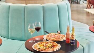 A dining area with a teal cushioned bench, two pizzas on plates, wine glasses, and bottles of Limonata and another beverage.