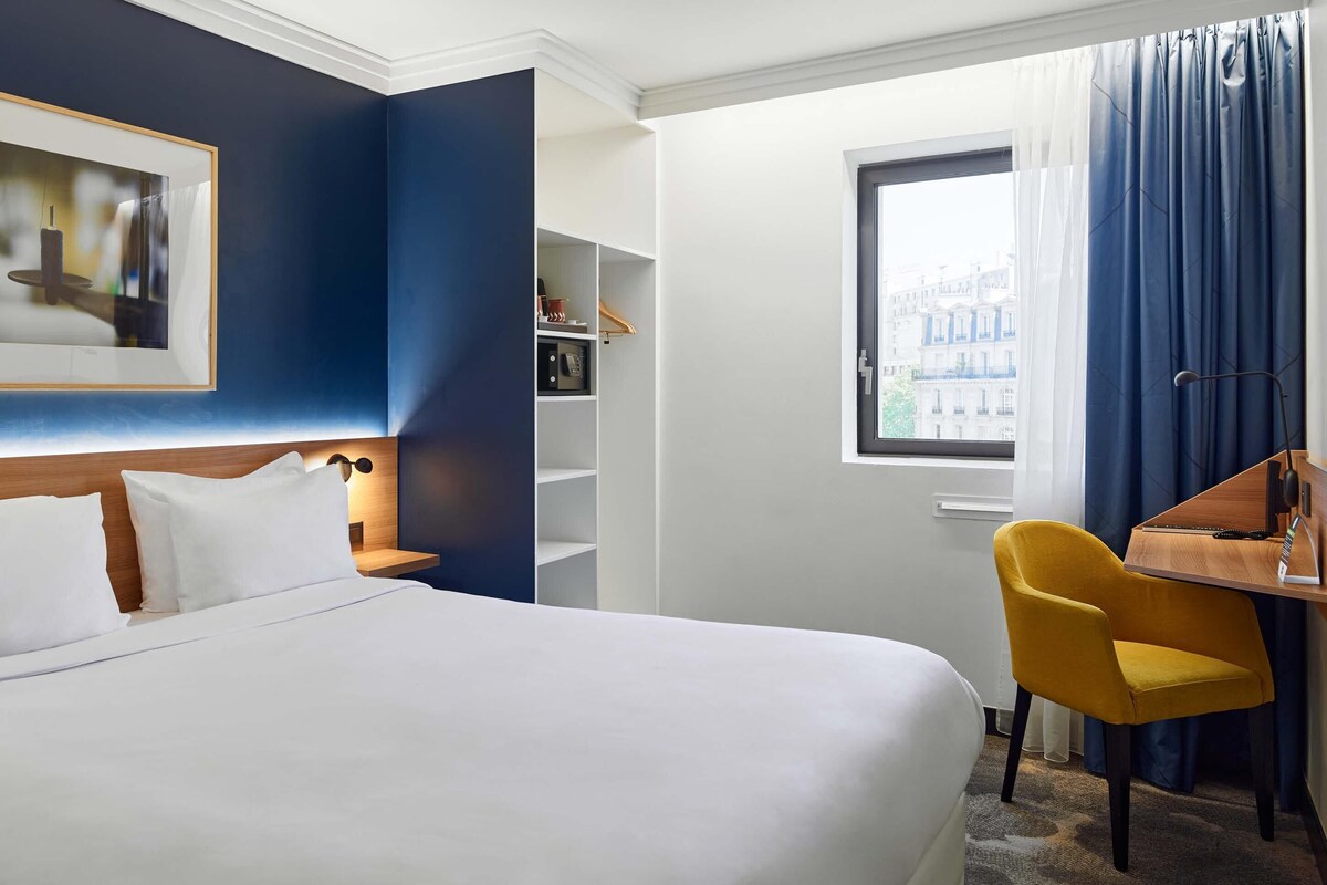 A hotel room with a bed, a desk, a chair, a wardrobe, and a window with curtains.