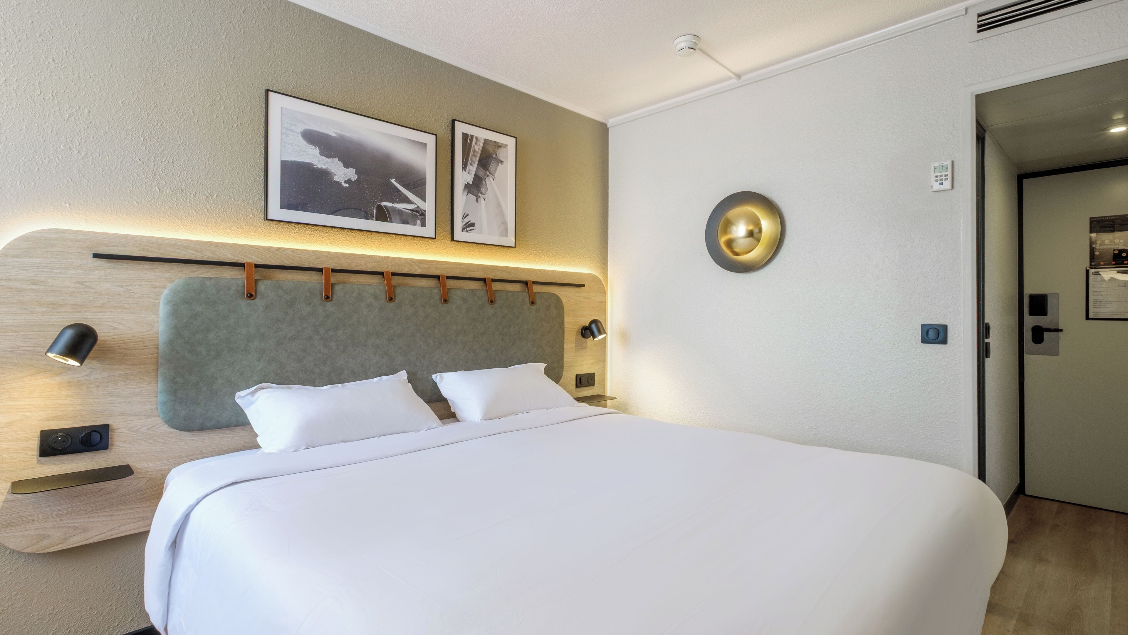 Quarto de hotel com uma cama grande, dois quadros fixados na parede, um relógio de parede redondo e uma mesinha de cabeceira com abajur.