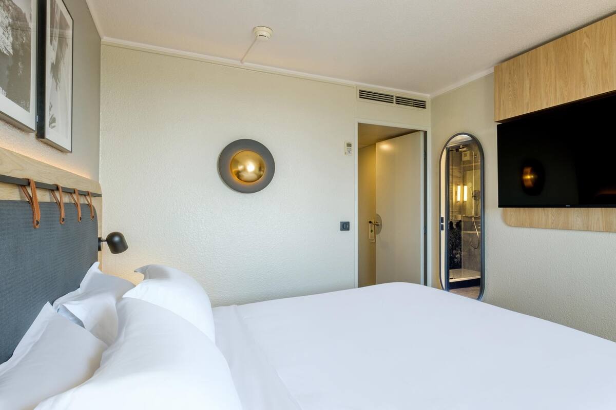 A hotel room with a bed, a mounted TV, a door leading to a bathroom, and a wall-mounted light fixture.