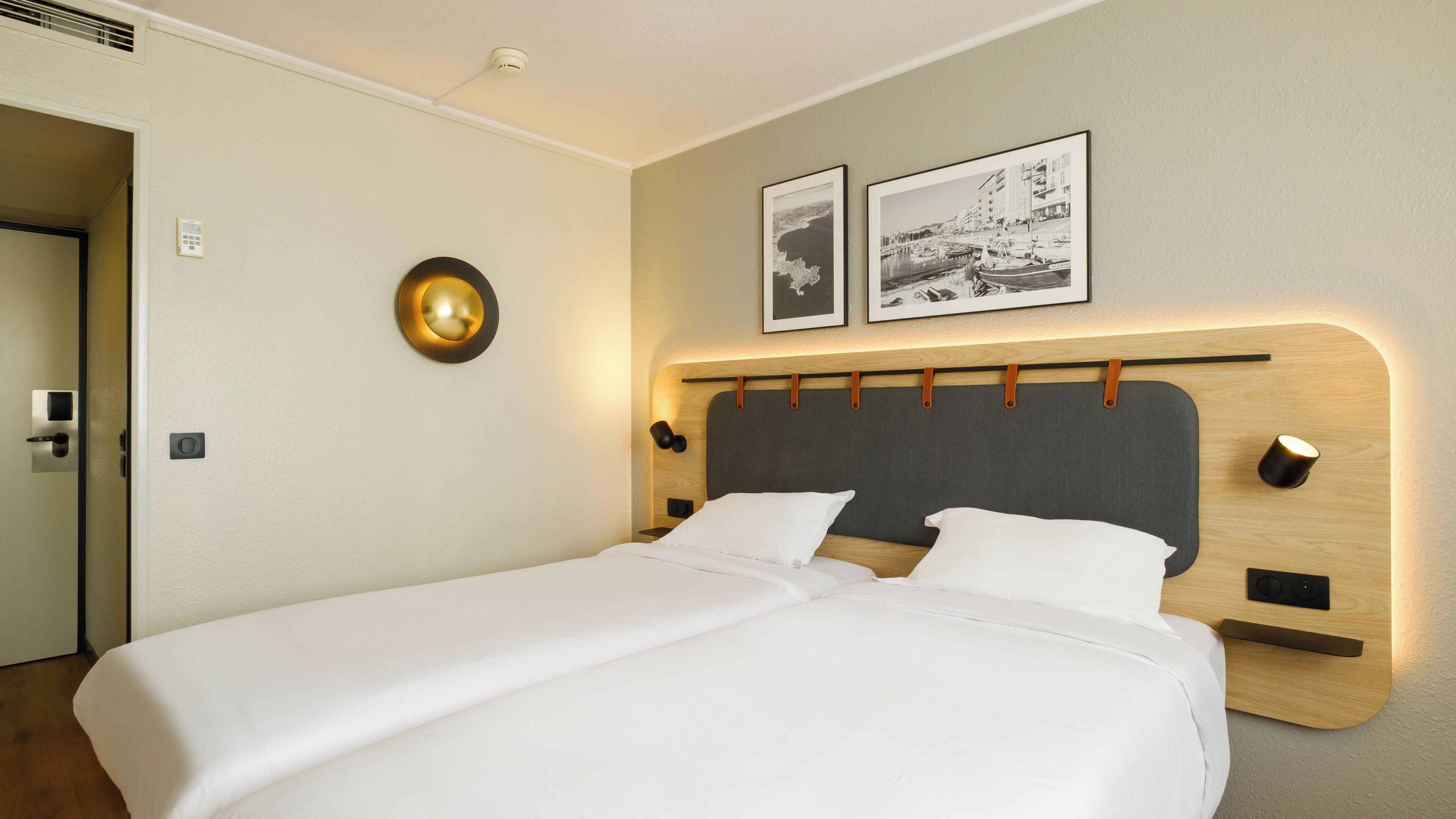 Une chambre d’hôtel avec un lit double, une lampe fixée au mur, deux tableaux encadrés et une porte avec une poignée.
