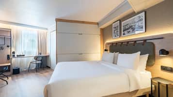 A modern hotel room with a large bed, a desk with a chair, and a wardrobe. There are two framed pictures on the wall above the bed.