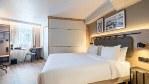 A modern hotel room with a large bed, a desk with a chair, and a wardrobe. There are two framed pictures on the wall above the bed. - Campanile PRIME - Nice Airport (Nice)