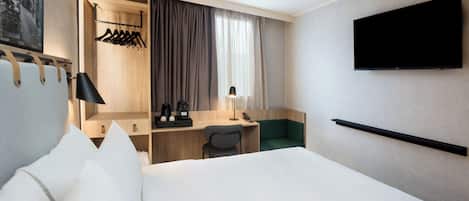 Quarto de hotel com cama, escrivaninha, cadeira, televisão e guarda-roupa com roupas penduradas.